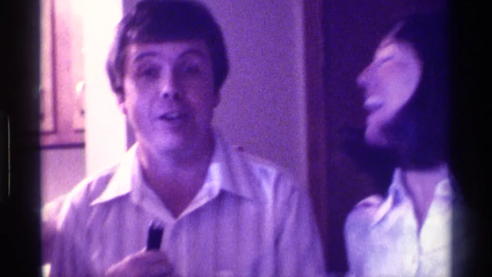 1976 Kentucky home-movie frame of a man with a drink near a laughing dark-haired woman in a dim indoor gathering.