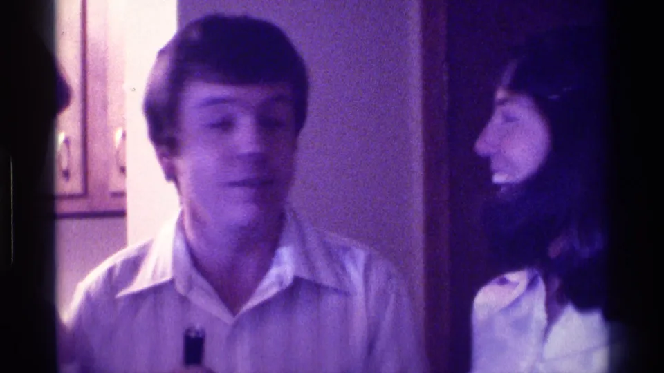 In 1976 Kentucky home-movie footage, a man in a light shirt stands near a woman in a white collared shirt holding a small black microphone.