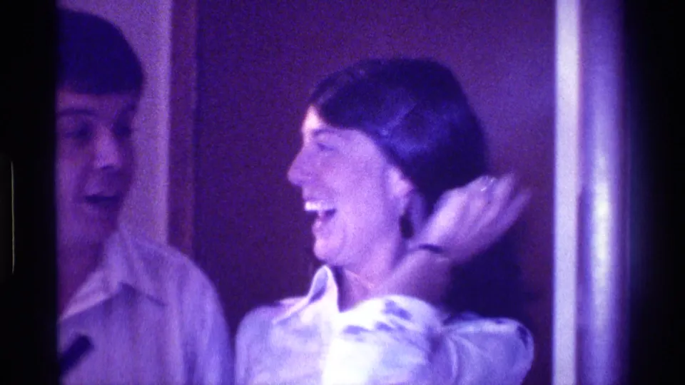 The woman in a white collared shirt laughs and turns toward the man beside her in this 1976 Kentucky indoor home-movie frame.