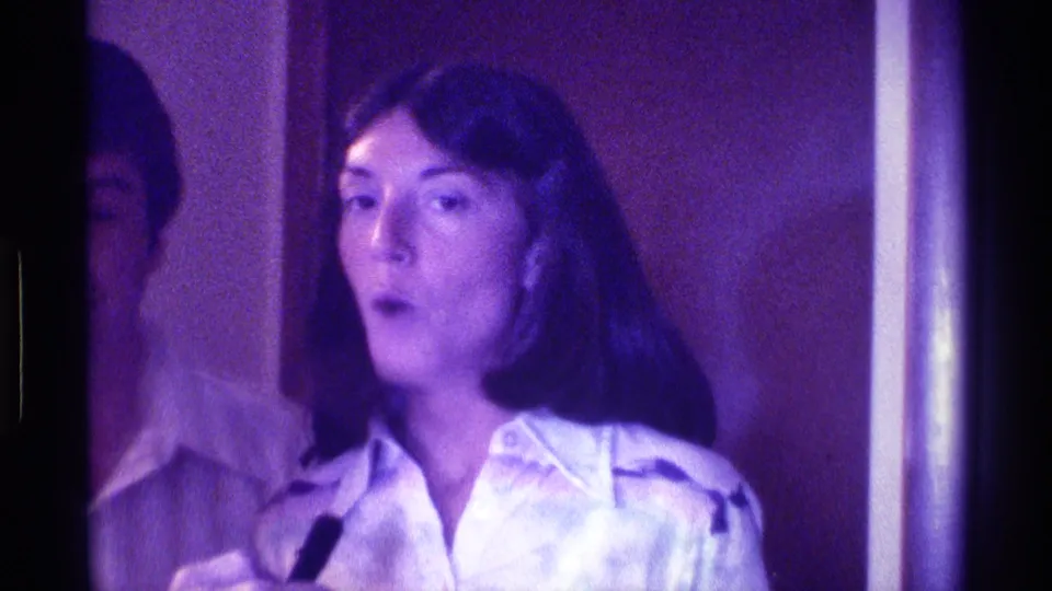 The woman faces forward with a small black microphone near her chest, framed by an interior wall and doorway in 1976 Kentucky footage.