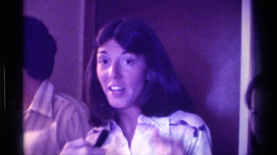 The woman gestures with one hand while holding the microphone, with a man partly visible beside her in the 1976 Kentucky home-movie scene.