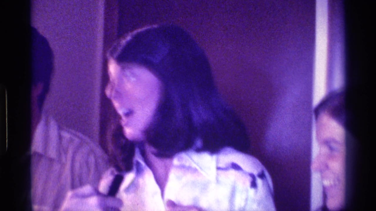 Woman in a white shirt holds a small black microphone beside partially visible people indoors.