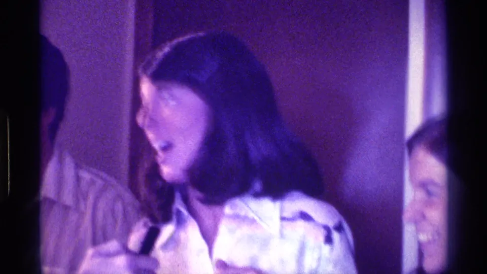 In 1976 Kentucky home-movie footage, a woman in a white shirt turns while holding a small black microphone in a wood-toned interior.