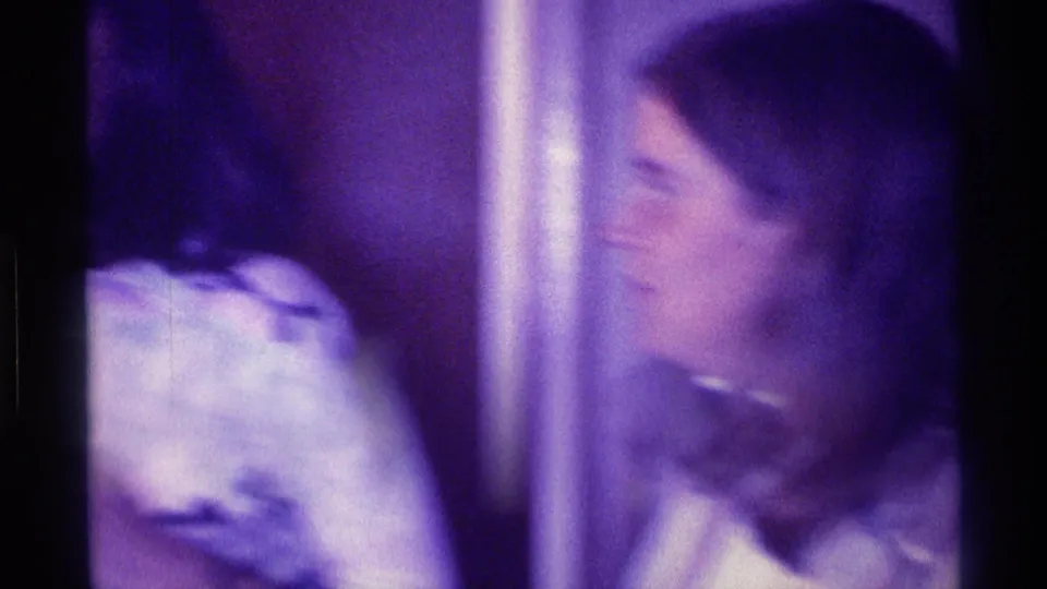 A blurred 8mm frame shows the woman and nearby people shifting in a Kentucky interior during the 1976 gathering.