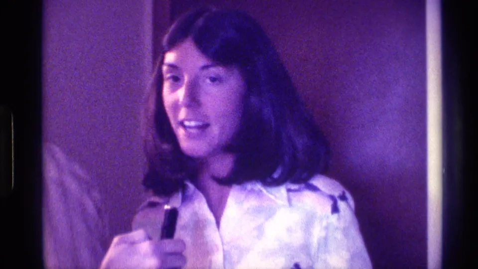 The woman faces the camera with a small black microphone in hand, with partial figures nearby in 1976 Kentucky home-movie footage.