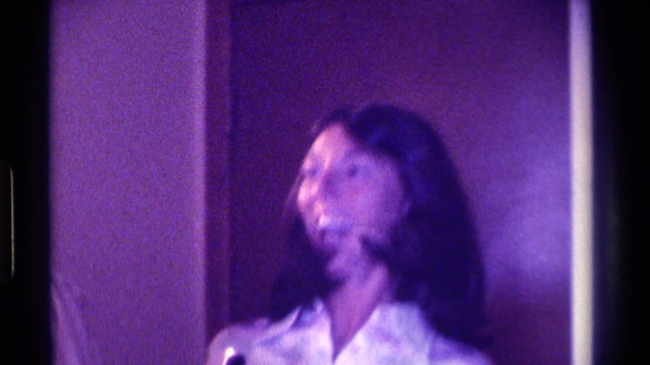 In 1976 Kentucky home-movie footage, a woman laughs while holding a microphone in a dim private interior with soft purple color shift.