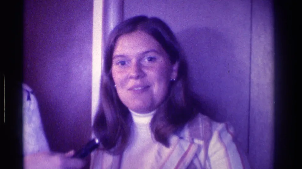 A woman in a turtleneck and light outer layer faces the camera in a dim 1976 Kentucky home-movie party setting.