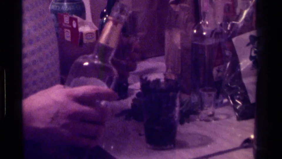 A party table in the 1976 Kentucky home movie shows bottles, cups, a pitcher, and food-or-drink items under dim purple-tinted light.