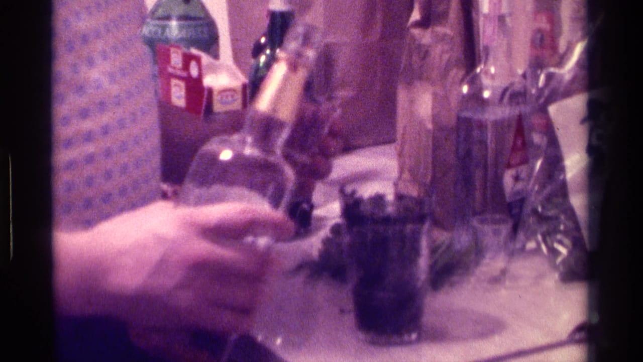 Hands and bottles on a table with a dark drink in a glass.