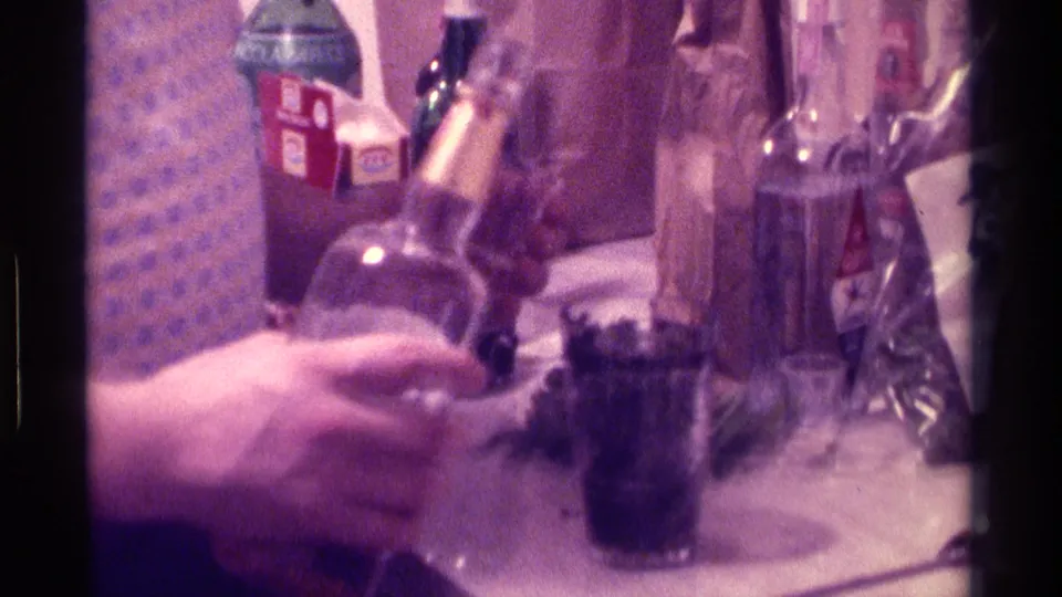 1976 Kentucky home-movie frame showing hands near liquor bottles, a tumbler with dark liquid, and a crowded indoor table.