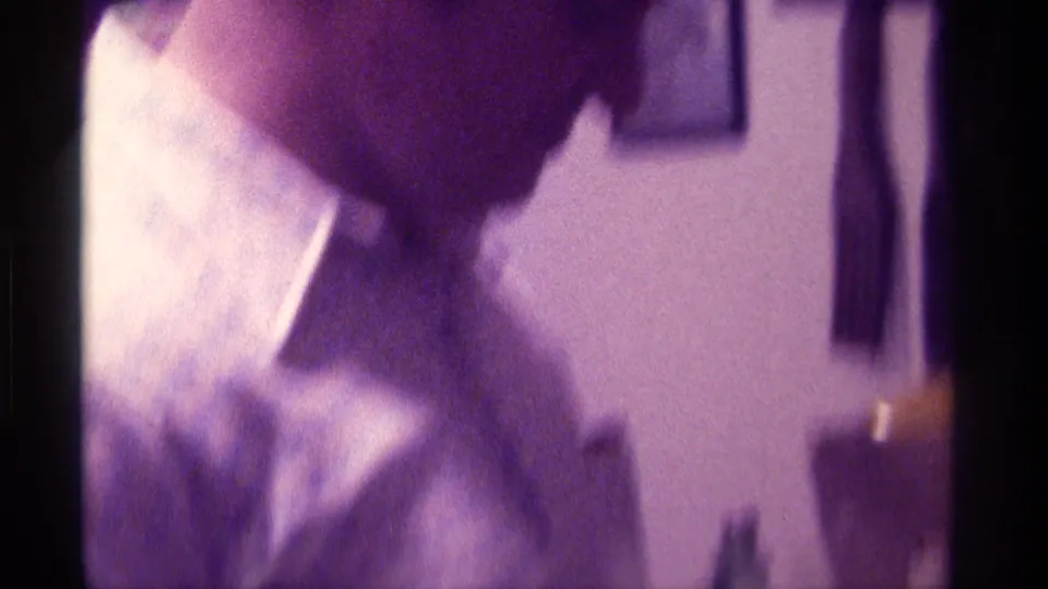 Soft, purple-shifted 8mm frame of a young man leaning forward inside a crowded Kentucky party room.