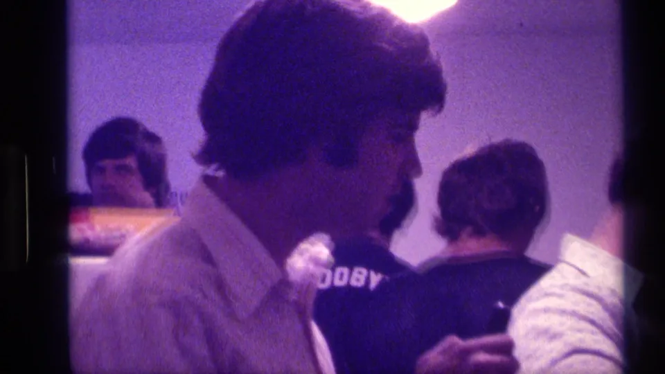 Grainy 8mm frame of several college-age men gathered in a dim Kentucky interior, with one face in profile near the camera.