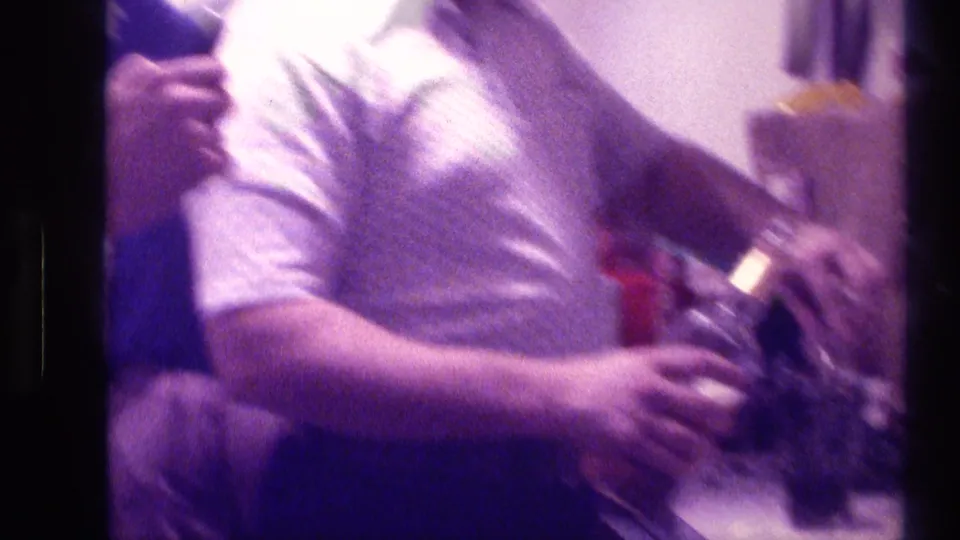 1976 Kentucky home-movie frame showing an adult man at a home party holding a bottle near a kitchen or bar counter.