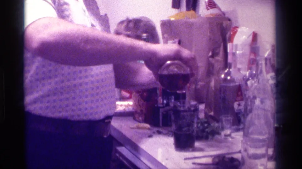 1976 Kentucky party footage showing a man in a patterned shirt pouring a drink among bottles, glasses, and a paper bag.