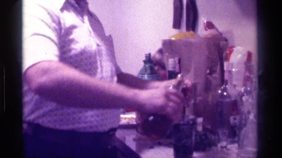 Close 1976 Kentucky home-party frame with hands handling a bottle at a kitchen or bar counter filled with bottles and glasses.