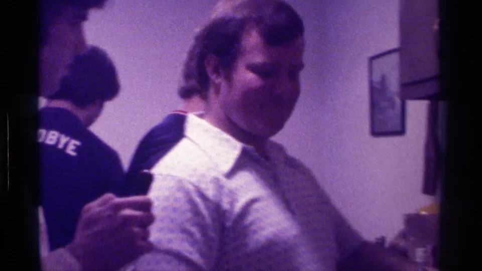 1976 Kentucky home-movie frame showing a man holding a drink while another guest stands behind him in a private party room.