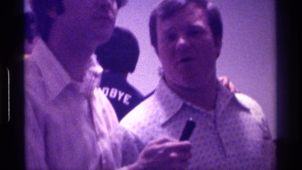 Two adult men indoors, one facing camera with a small dark object, with a GOODBYE shirt visible behind them.