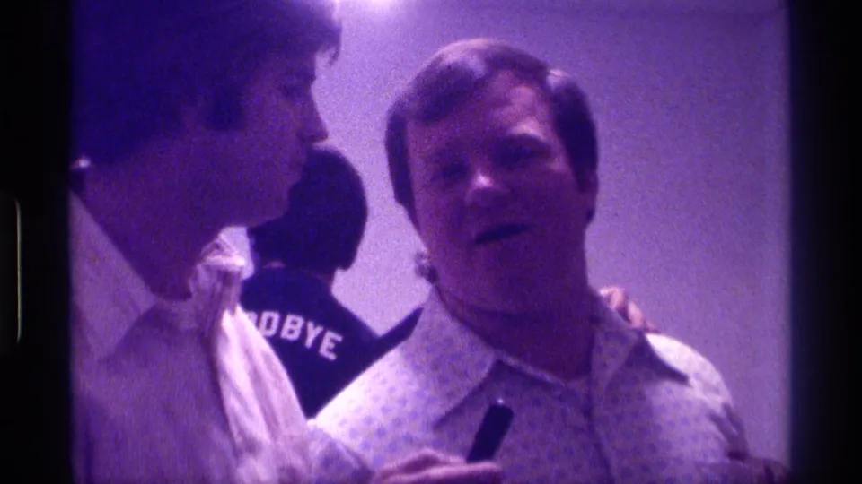 1976 Kentucky home-movie frame showing a close indoor conversation between two adult men, with readable GOODBYE text on a dark shirt in the background.