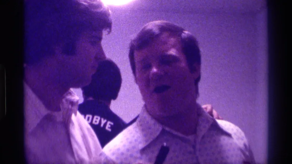 1976 Kentucky 8mm still of two adult men in a tight indoor home-movie composition, one speaking while a person in a GOODBYE shirt stands behind.