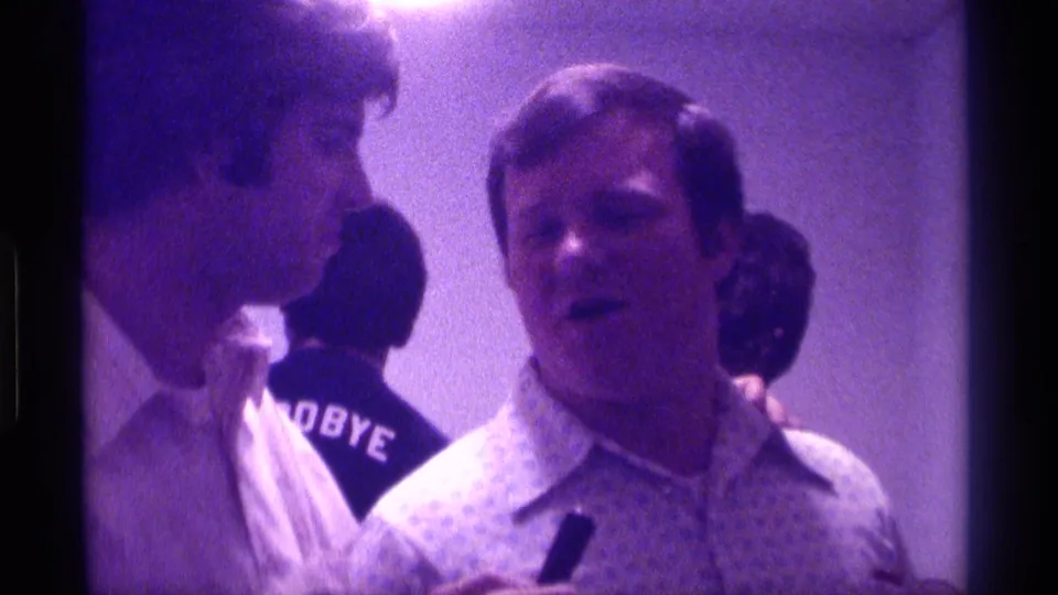 1976 Kentucky home-movie frame of an informal indoor interview moment, showing two adult men in close view and a readable GOODBYE shirt in the background.