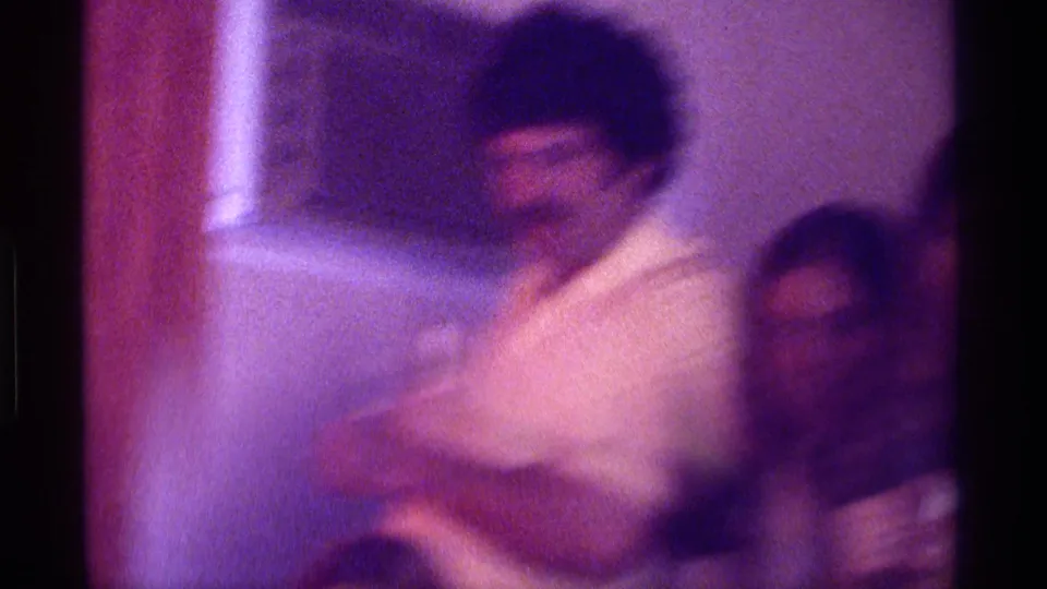 Kentucky 1976 home-movie frame showing a blurred man in a light shirt near a doorway or wall, with another adult partly visible.