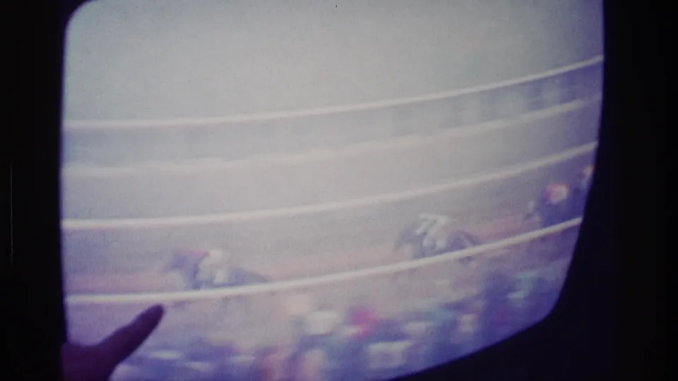 1976 Kentucky home-movie frame with a hand pointing at a CRT screen as horses race behind pale track rails.