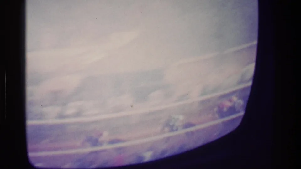 Color-faded 8mm frame of a television race broadcast, with the track and riders barely visible through soft exposure.