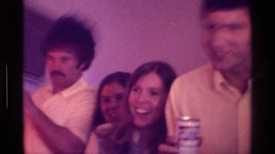 1976 Kentucky home-movie frame of adults smiling and cheering in a private living room while watching television.