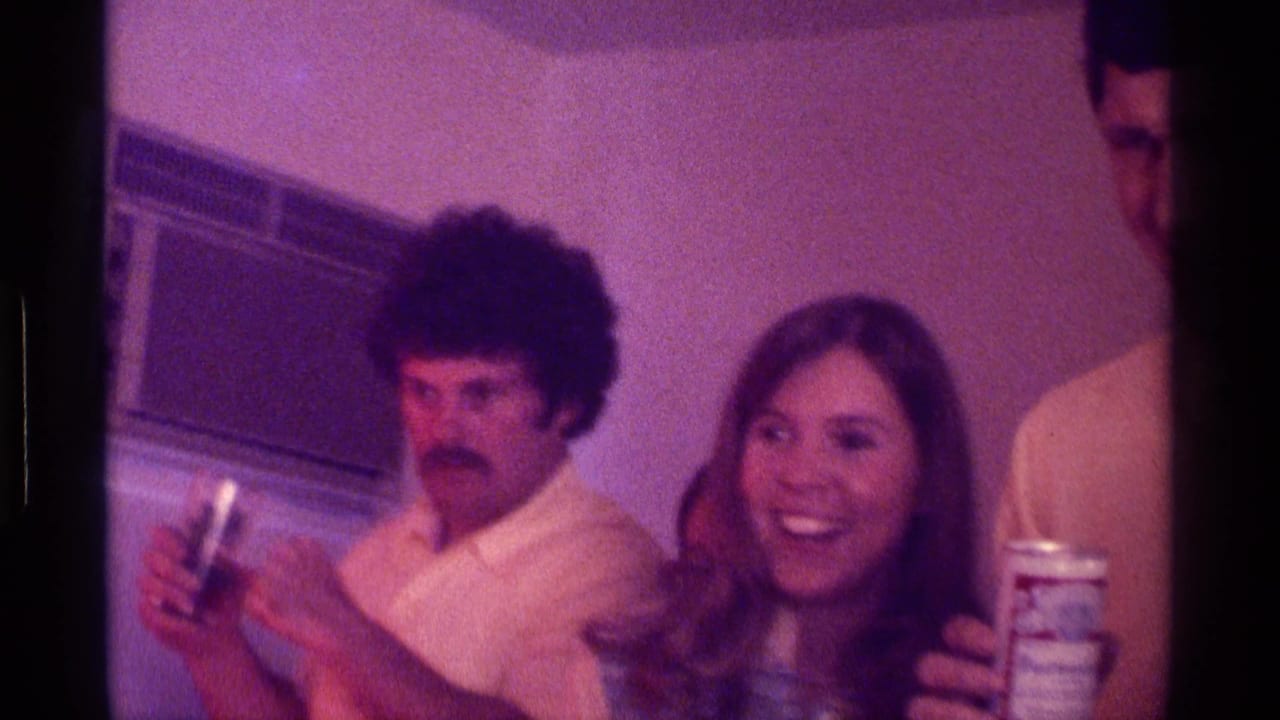 Young adults indoors at a party, with a man holding a glass and a woman smiling near beverage cans.