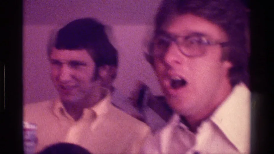1976 Kentucky party footage frame with men in collared shirts, one in eyeglasses reacting near the handheld camera.