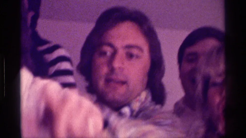 1976 Kentucky home-movie close-up of a young man at an indoor party, with other faces and a striped sleeve nearby.
