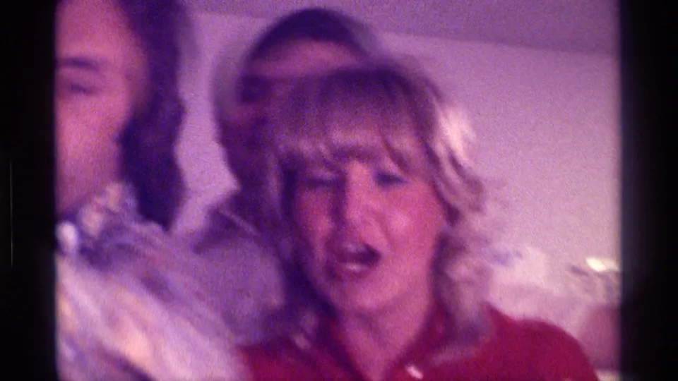 1976 Kentucky home-movie frame of a woman in a red top reacting animatedly among friends in a private interior.