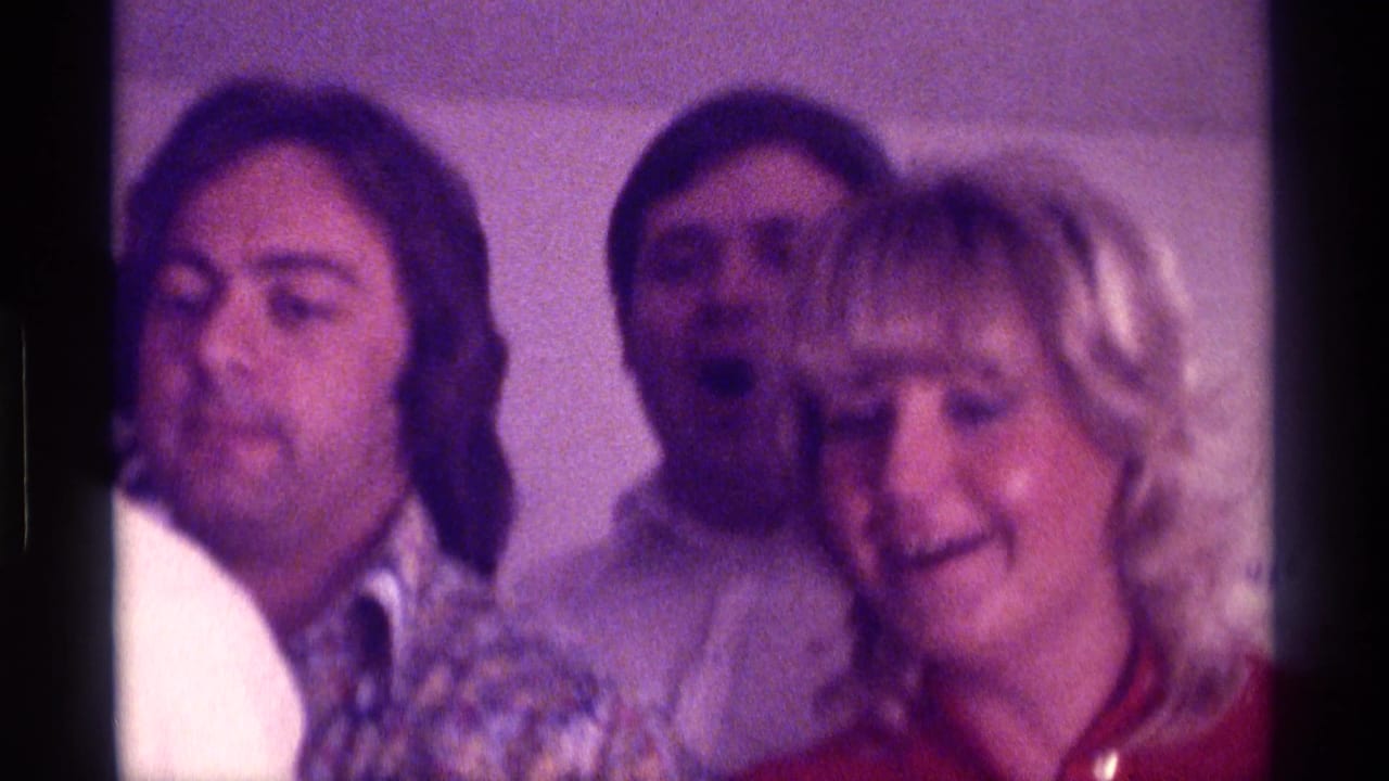 Adults crowded together at an indoor party, including a blonde woman and men in patterned shirts.
