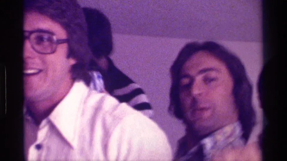 A smiling adult man in large glasses and a wide-collar white shirt turns toward the camera at a 1976 Kentucky indoor party.