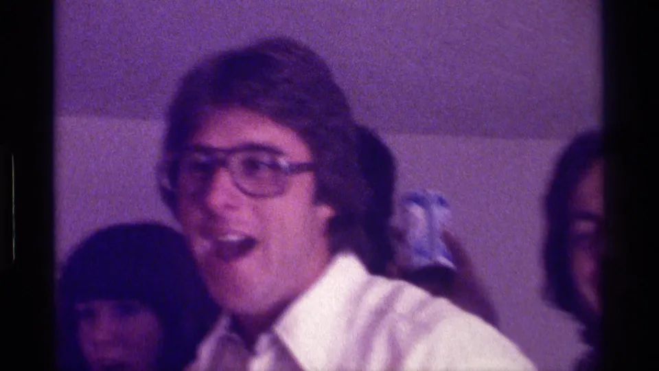 Close 8mm frame of a smiling man in large glasses at a Kentucky party, surrounded by partially visible adult guests.