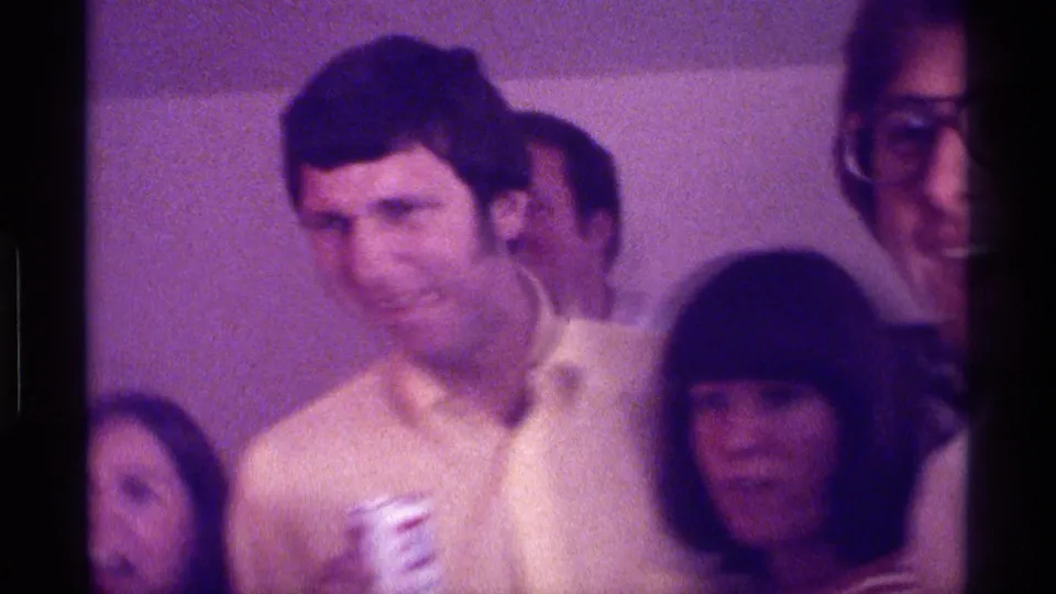 1976 Kentucky party frame showing a man in a pale yellow shirt holding a cup, with adult guests seated close around him.