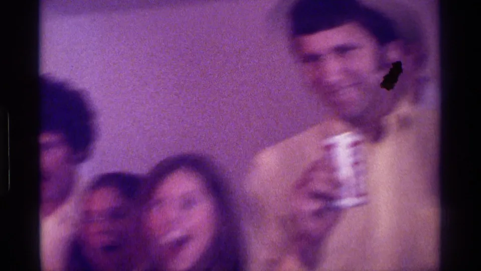 1976 Kentucky home-movie frame showing adults in a dim room, with a man in a light shirt holding a beer can as friends react nearby.
