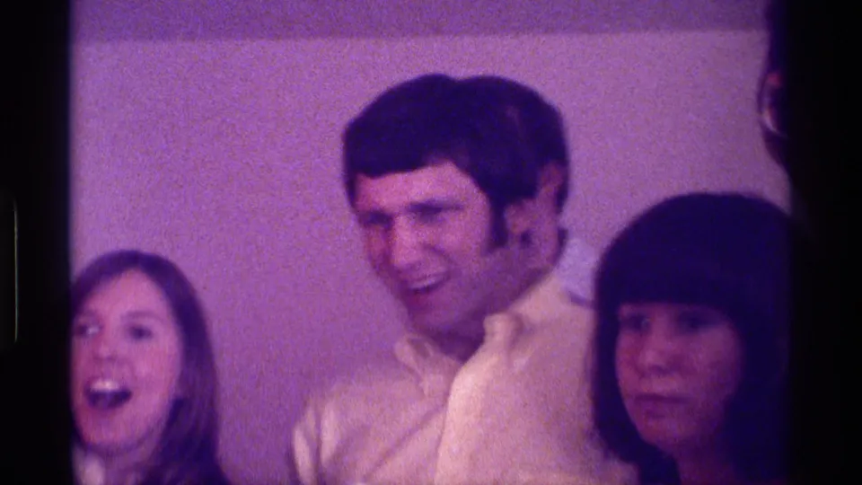1976 Kentucky home-movie frame with a smiling man in a light collared shirt between women at a private indoor gathering.
