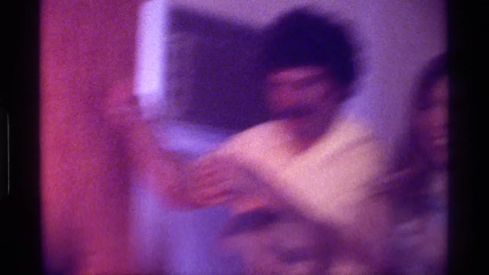 Soft, blurred 1976 Kentucky home-movie frame of a man moving in a dim indoor gathering, with another face partly visible at the edge.