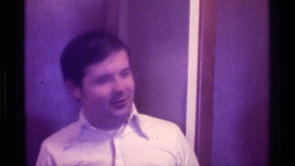 The man faces slightly left in the interior setting, with a white shirt, dark hair, wall trim, and strong purple 8mm color shift visible.