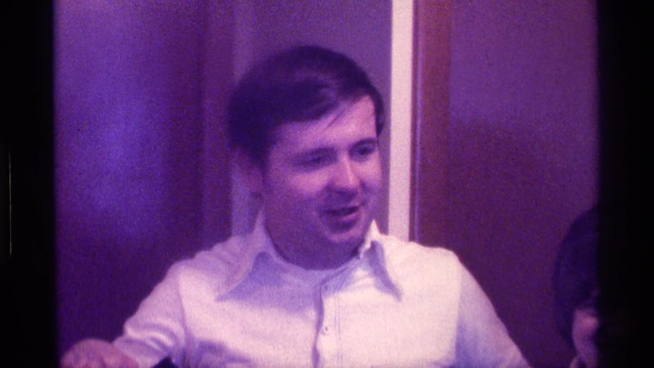 Adult man in a white collared shirt stands indoors beside a wood wall frame.