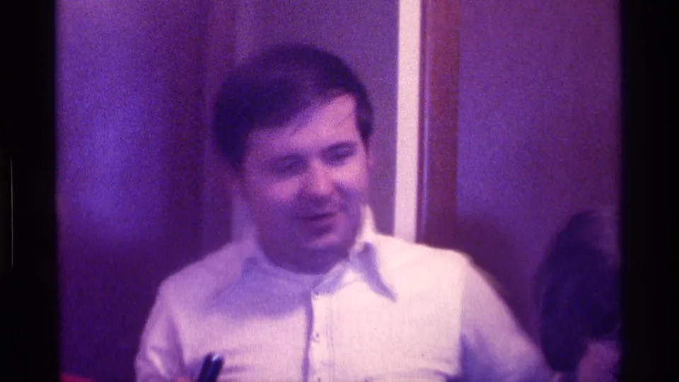The man remains near the wood-paneled wall in the 1976 Kentucky interior, with a partial nearby face at the right edge.