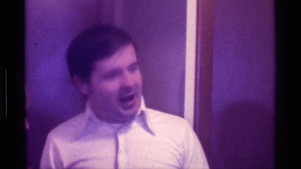 In the purple-tinted 8mm frame, the adult man appears animated or mid-speech beside a wood-paneled wall in Kentucky.