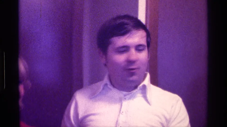 The man stands near the wood wall frame in the 1976 Kentucky home movie, with another person's partial face visible at the left edge.