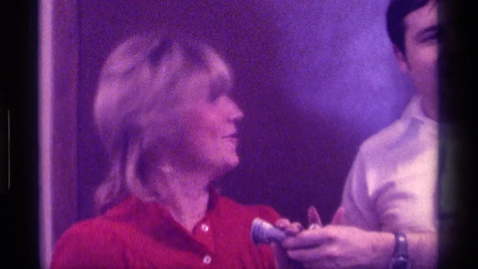 1976 Kentucky home-movie frame of a blonde woman in red looking toward a man in a white shirt holding cash or a small item.