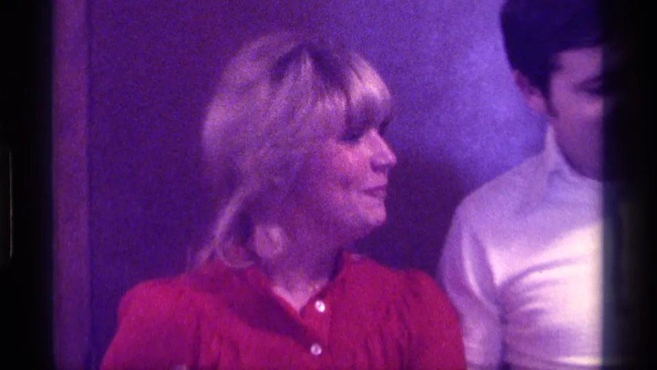 1976 Kentucky 8mm frame showing the blonde woman in red in profile, with a man partly visible beside her indoors.