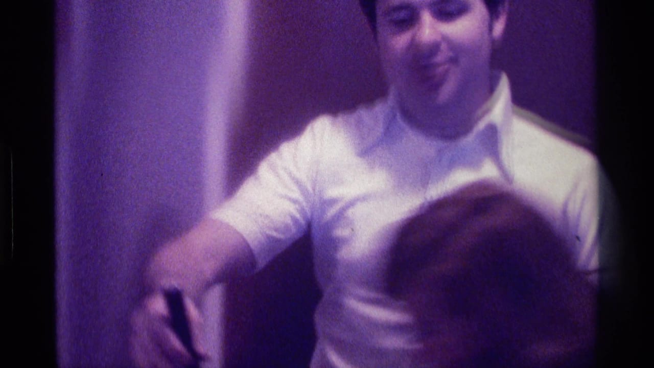 Man in a white collared shirt smiles indoors beside a partially visible seated person.
