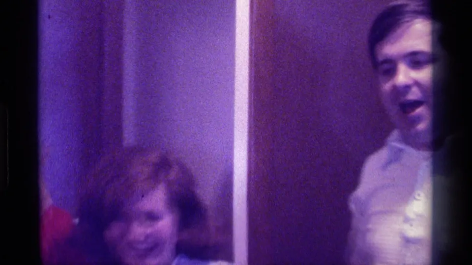 A smiling woman in a plaid jacket appears seated while a man stands nearby in a dim 1976 Kentucky indoor home-movie frame.