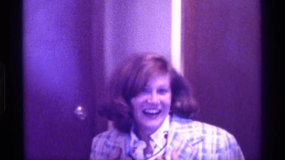 The woman in a plaid jacket laughs toward the camera in a softly focused 1976 Kentucky home-movie interior.