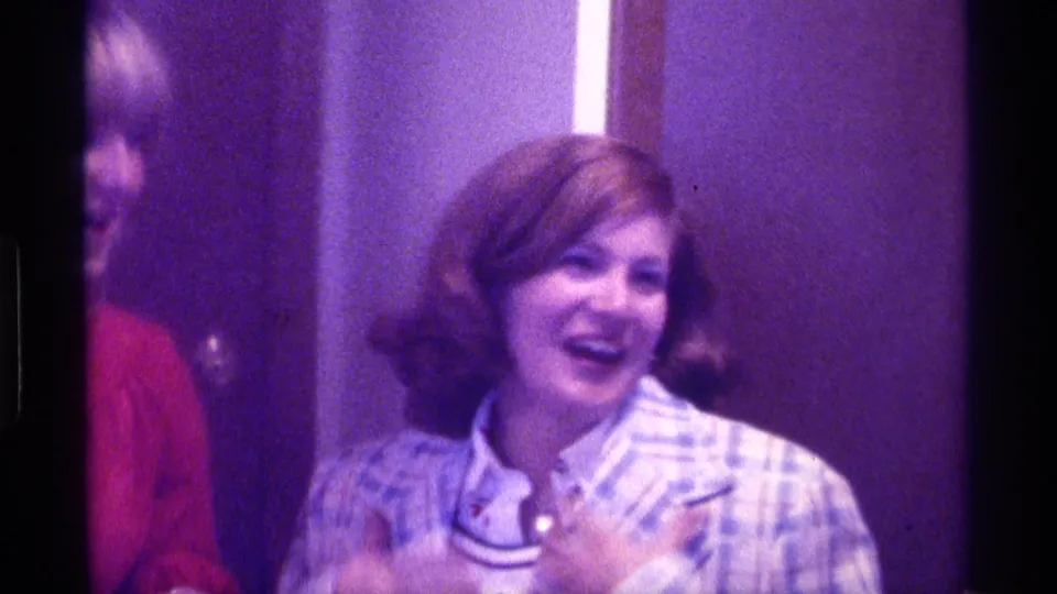 A smiling woman in a plaid jacket gestures toward her collar as another person is partly visible at the edge of the 1976 Kentucky interior frame.
